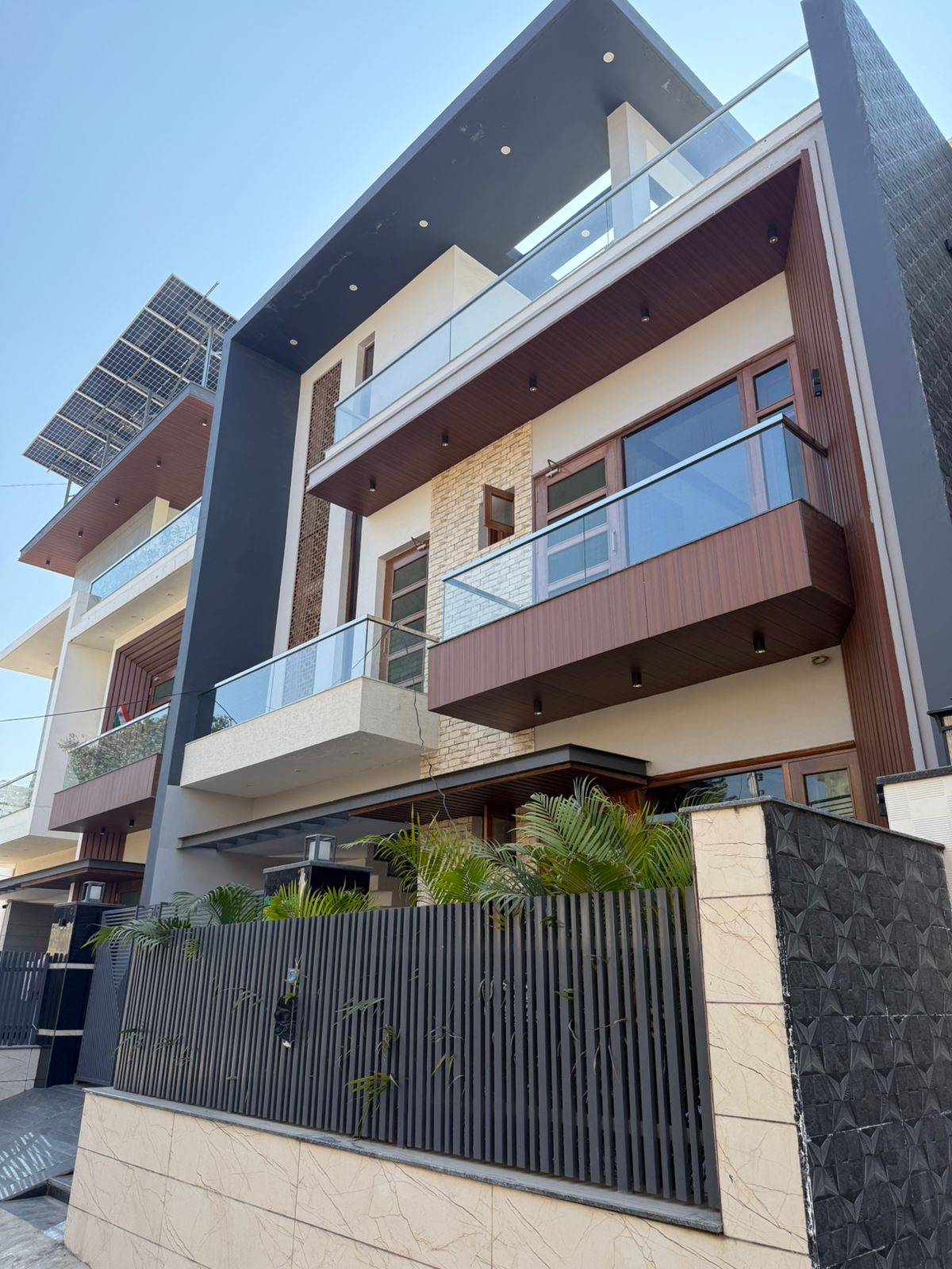 Modern three-storey luxury house with glass balcony railings, wooden cladding, stone accent wall, and black vertical facade elements.